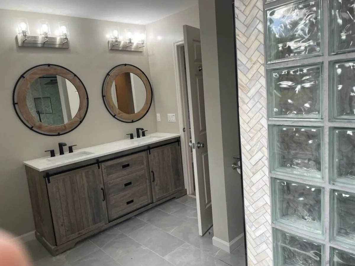A modern bathroom featuring a double vanity with two round mirrors, sleek light fixtures, and a glass block shower wall. Morning sunlight streams through, creating an airy atmosphere ideal for self-care and preparation.