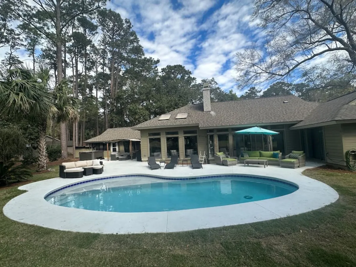 The hero image showcases a sun-drenched swimming pool with a spacious tanning shelf, surrounded by chic outdoor seating—including a plush green sofa and a stylish umbrella—nestled among tall, swaying trees. The space invites guests to swim, lounge, dine al fresco, or simply unwind in the shade, all within a lush, private setting.