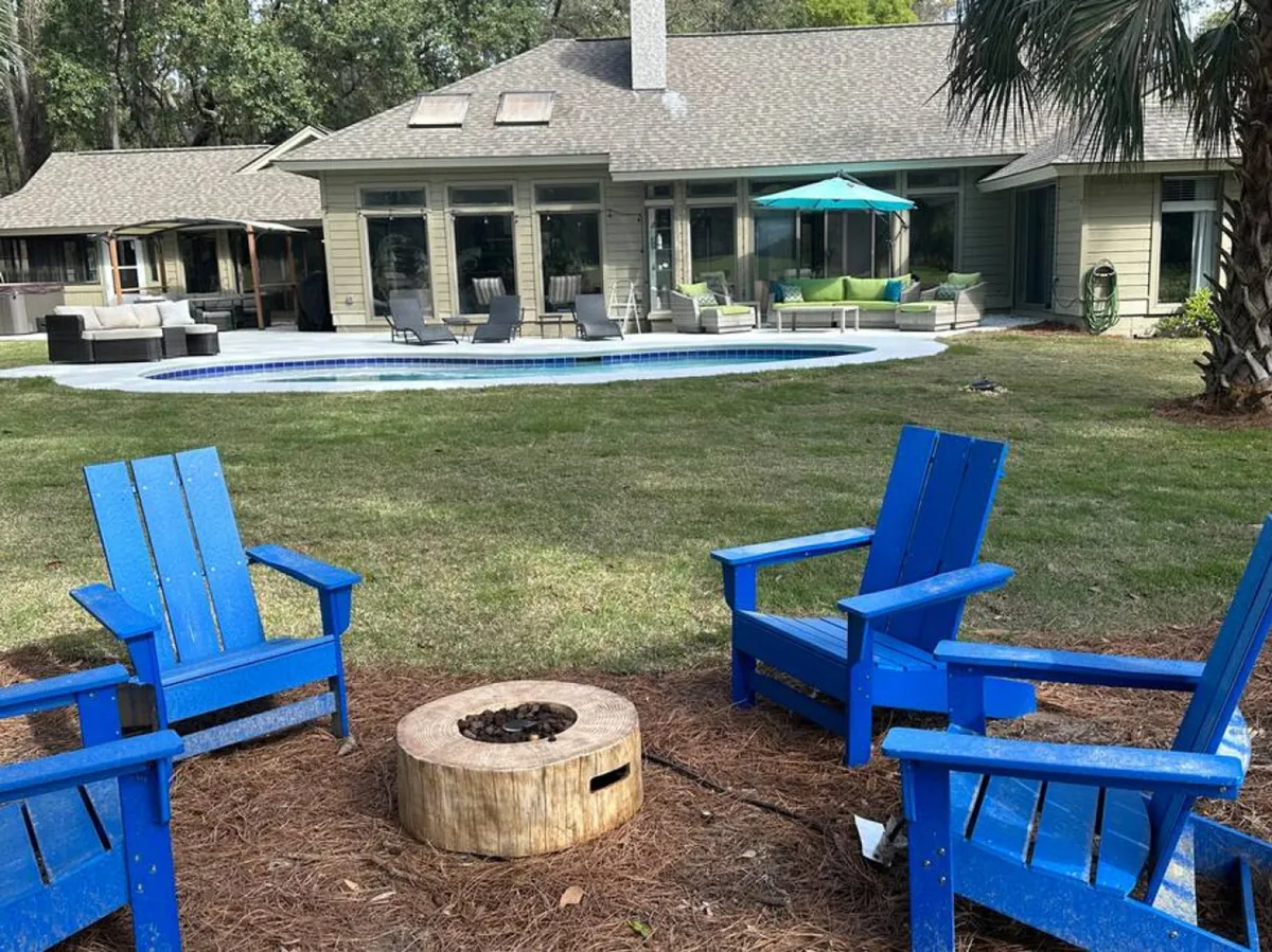 This vibrant exterior image showcases blue Adirondack chairs arranged around a glowing fire pit, with a sparkling swimming pool and multiple outdoor seating areas in view. The scene suggests a lively yet inviting space for both relaxation and social gatherings. Lush landscaping surrounds the area, enhancing the resort-like atmosphere.