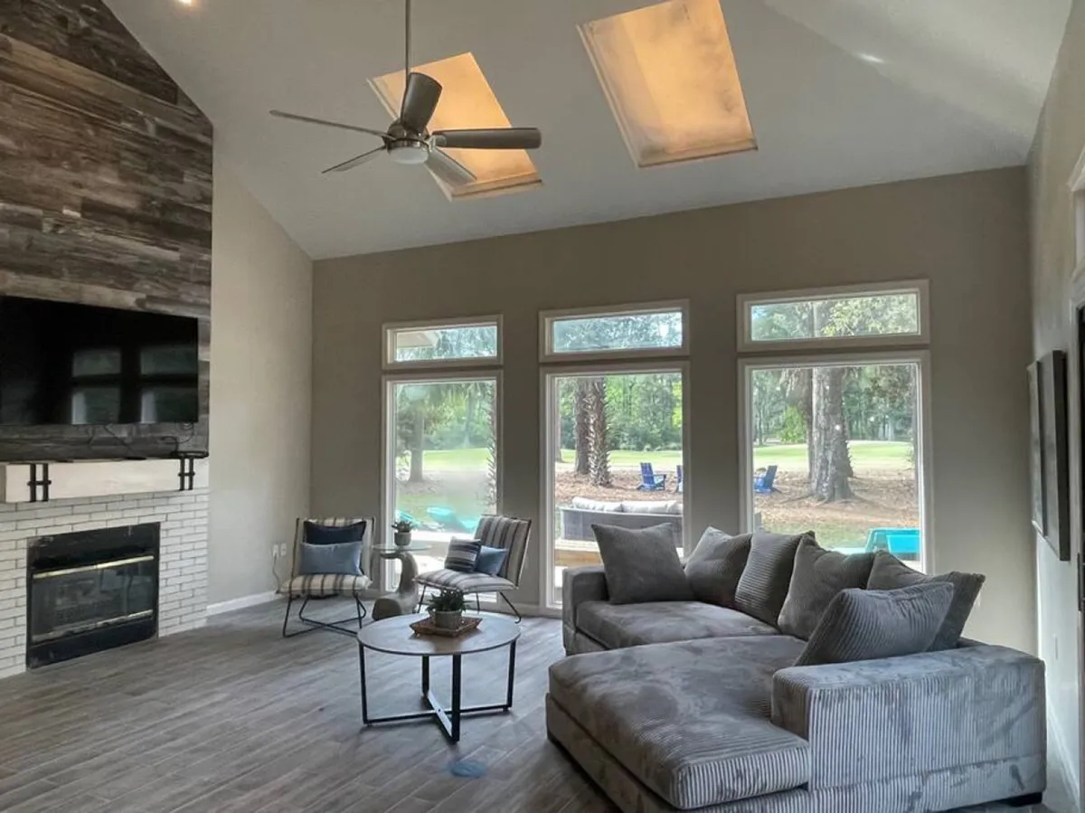A modern, airy living space with a large sectional sofa, a prominent fireplace, expansive windows offering lush outdoor views, and a ceiling fan overhead, creating an inviting atmosphere for relaxation and entertaining.