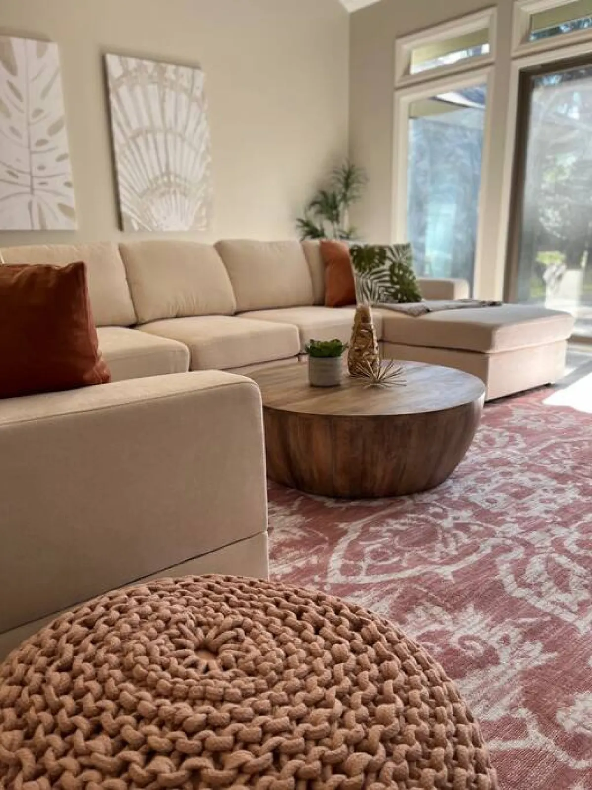 A welcoming living area with a stylish sofa and coffee table, decorated with tasteful wall art, a leafy plant, and a knitted pouf, radiating warmth and contemporary comfort for guests to socialize or unwind.