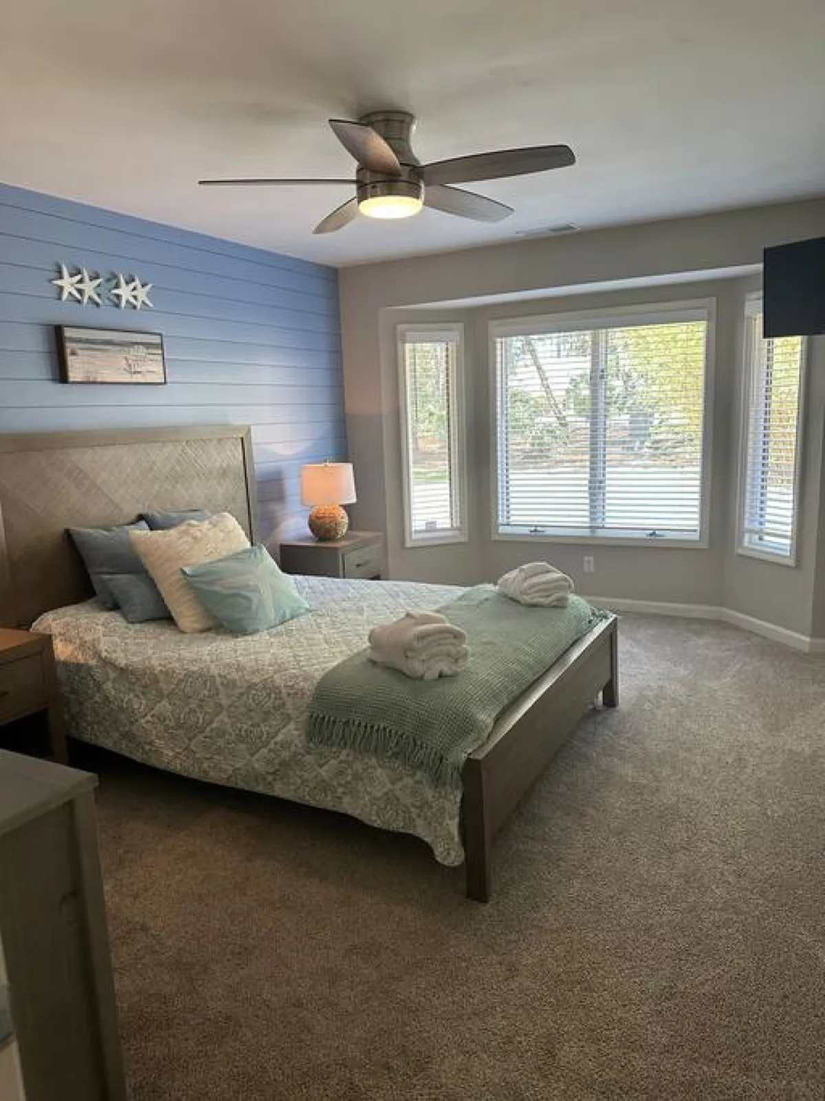 A sunlit bedroom with a comfortable bed, expansive bay windows overlooking the outdoors, a ceiling fan, and whimsical decorative stars. The setting exudes warmth and offers a serene spot for relaxing, reading, or enjoying the view outside.