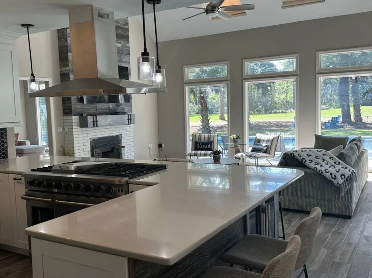 An open, sunlit kitchen and seating area featuring a large island, stainless steel appliances, and expansive windows that frame the outdoors. The space is designed for gathering, cooking, and entertaining.