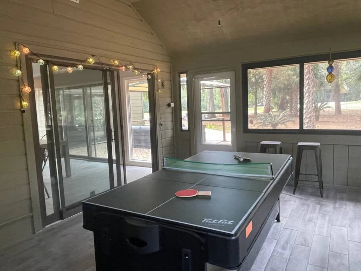 This image showcases a lively game room featuring a ping pong table lit by cheerful string lights. Large windows flood the space with natural light, while the setup encourages social interaction and friendly competition. The overall atmosphere is playful and inviting, ideal for all ages to unwind and connect.